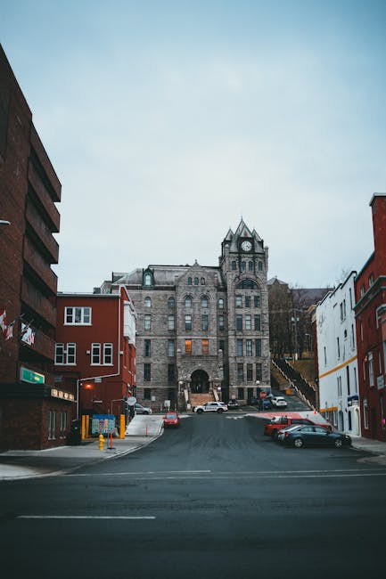 A wide-view street scene in a city showing a steep incline leading up to an old stone building with a clock tower and arched entrance, surrounded by colorful red and white buildings on either side. Th
